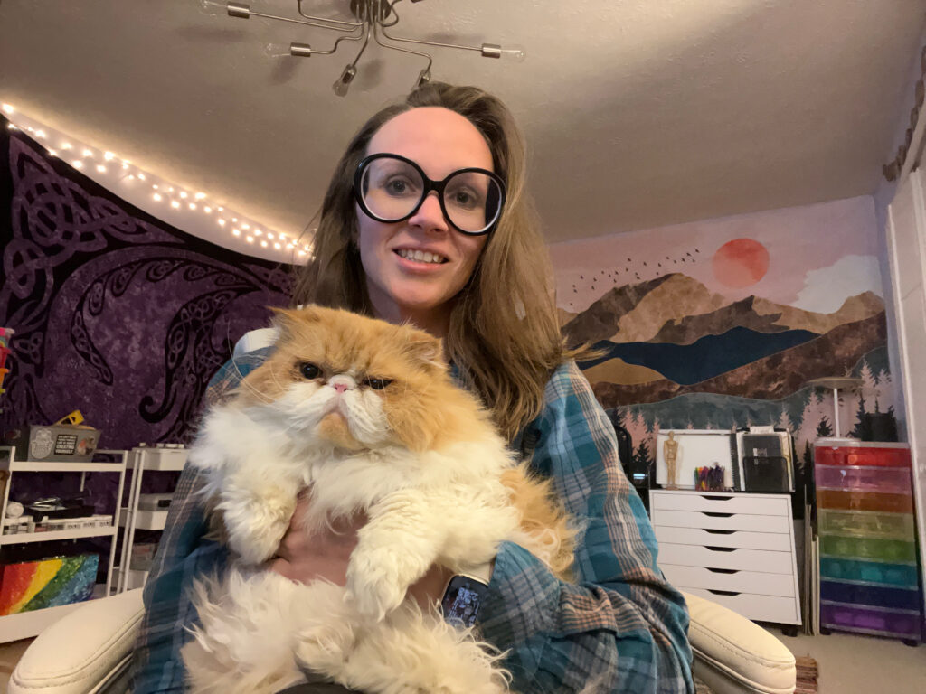 Jenna Stone with long brown hair and oversized black glasses smiles wearing a flannel while holding a fluffy orange-and-white Persian cat. They are seated in a cozy, colorful office with a mountain mural, purple tapestry, and rainbow storage drawers.